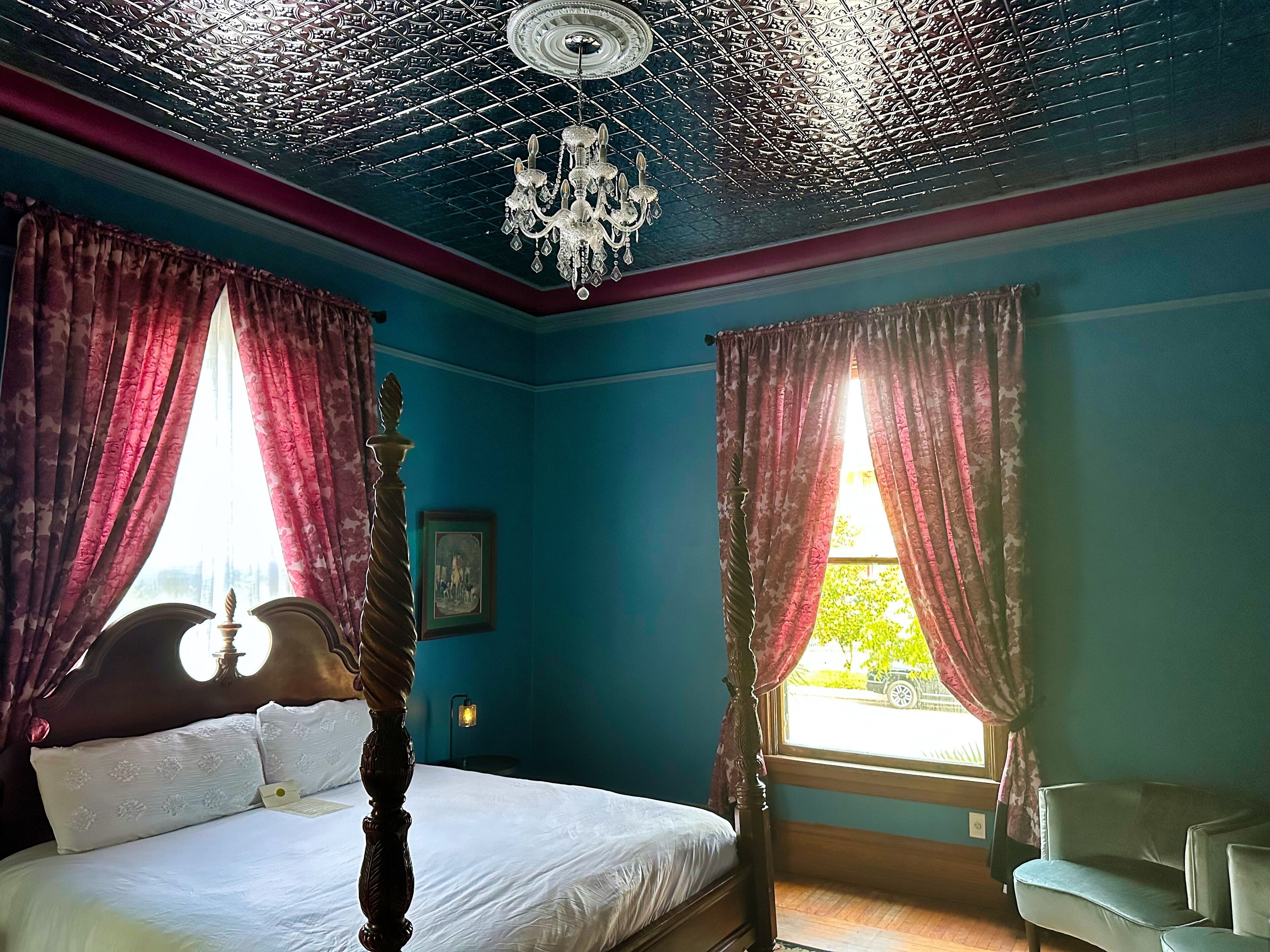 A bedroom with a four-poster bed, a chandelier, and a view of the outdoors through a window with curtains.