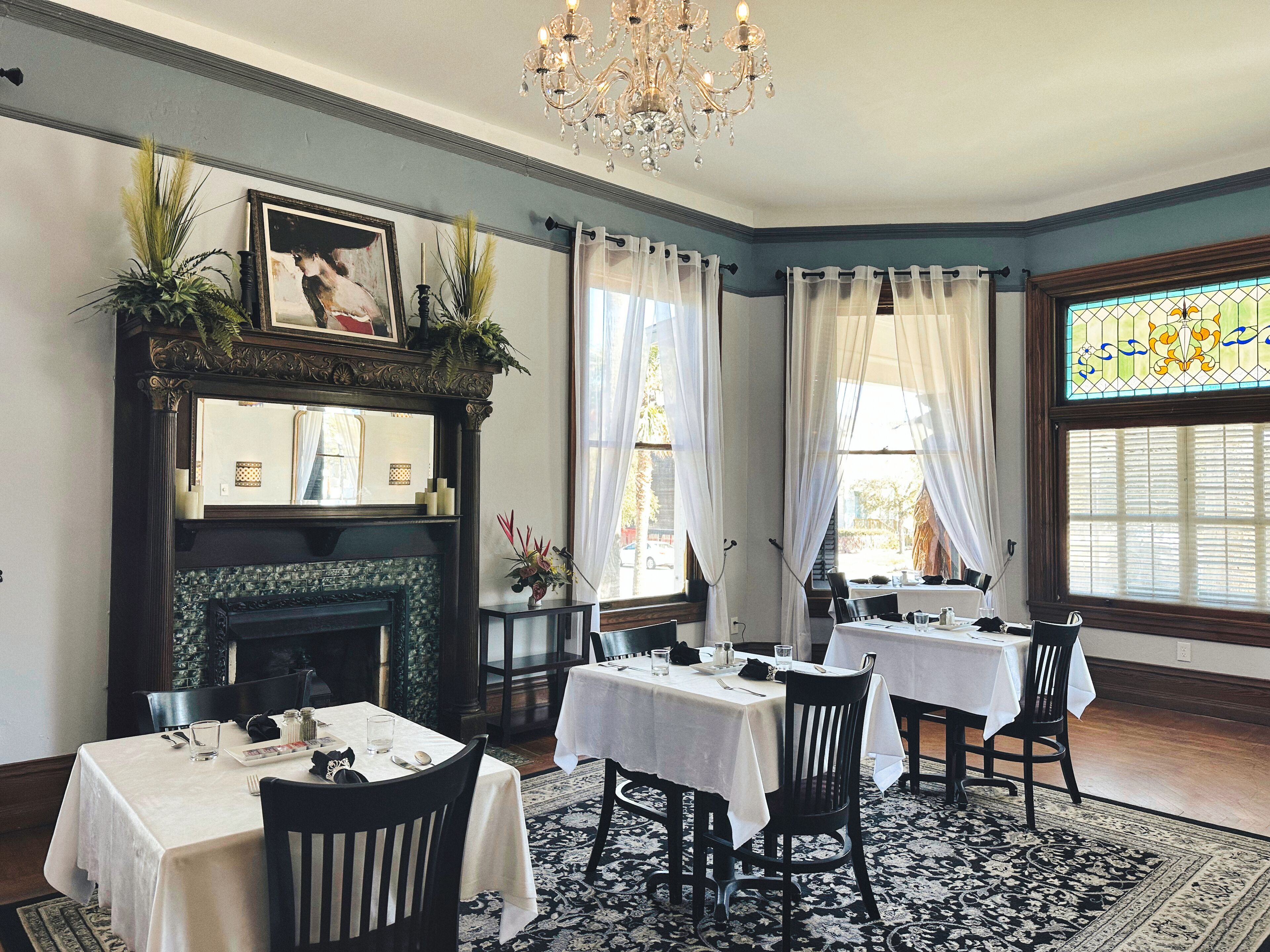 Uma sala de jantar com mesas preparadas para dois, uma lareira com espelho acima e um candelabro.