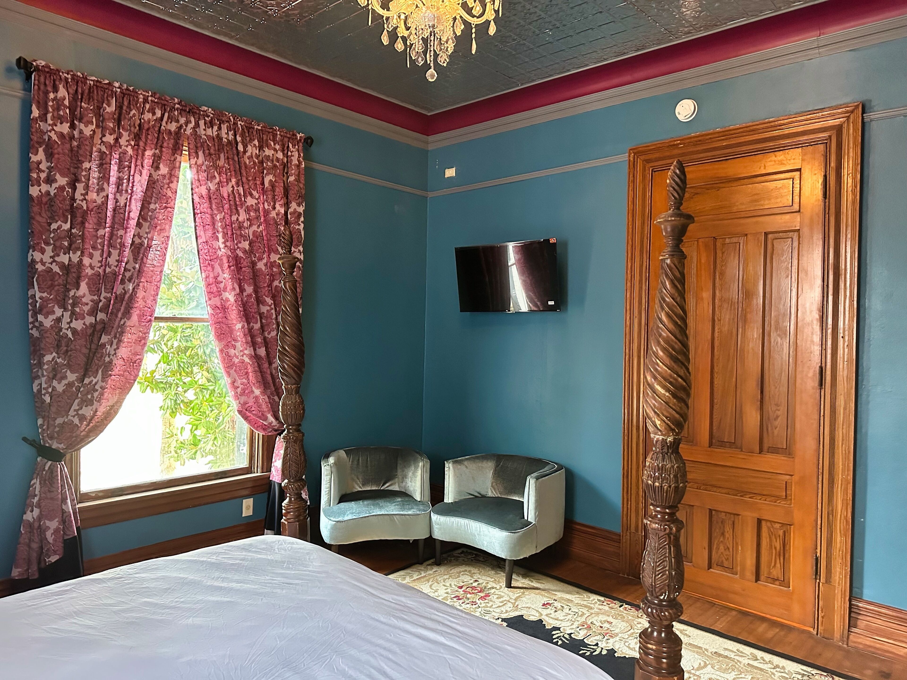 A bedroom with a four-poster bed, a television mounted on the wall, a wooden door, and two armchairs.