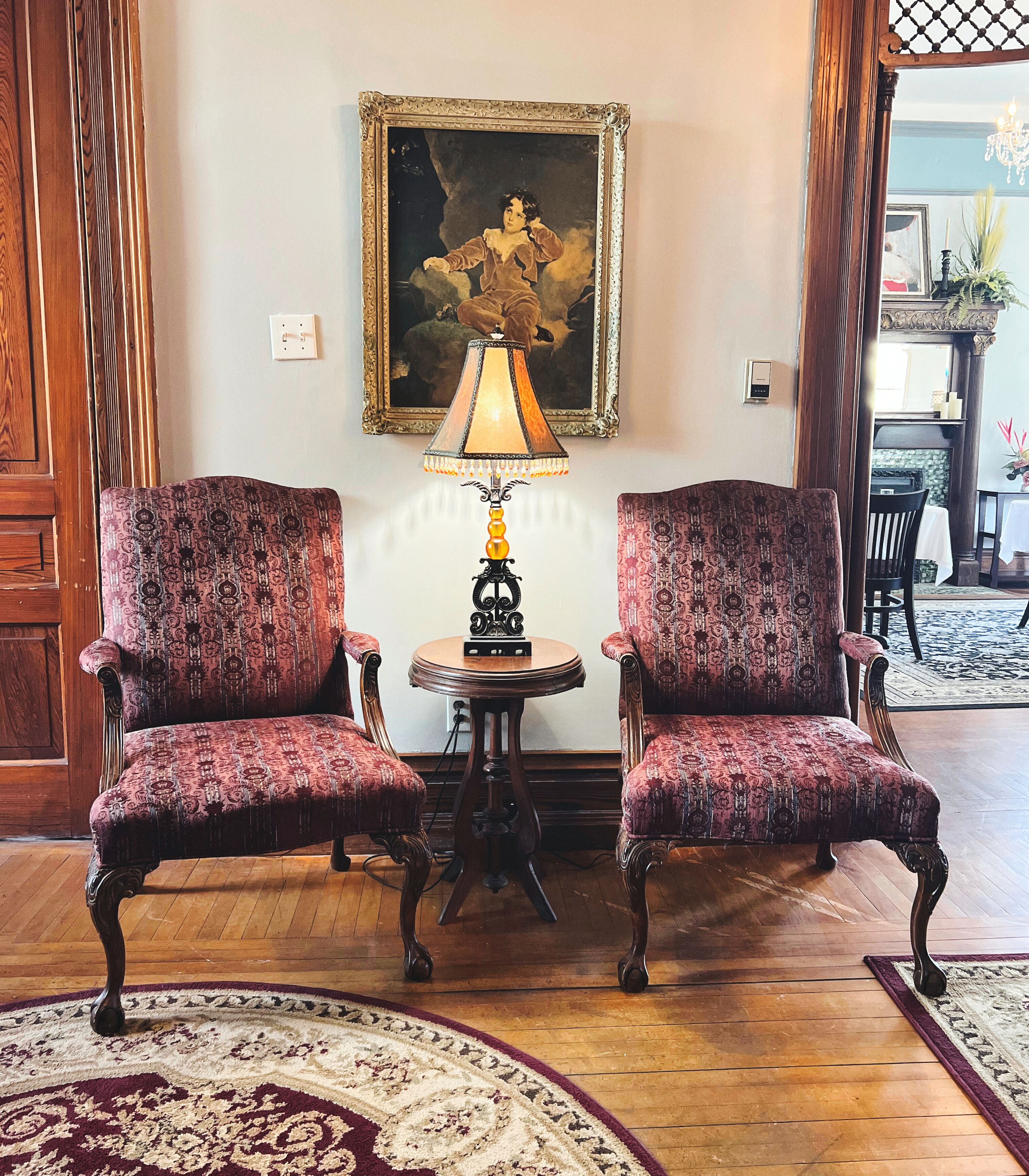 A room with two ornate chairs, a table with a lamp, and a framed painting on the wall.