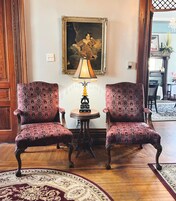A room with two ornate chairs, a table with a lamp, and a framed painting on the wall.