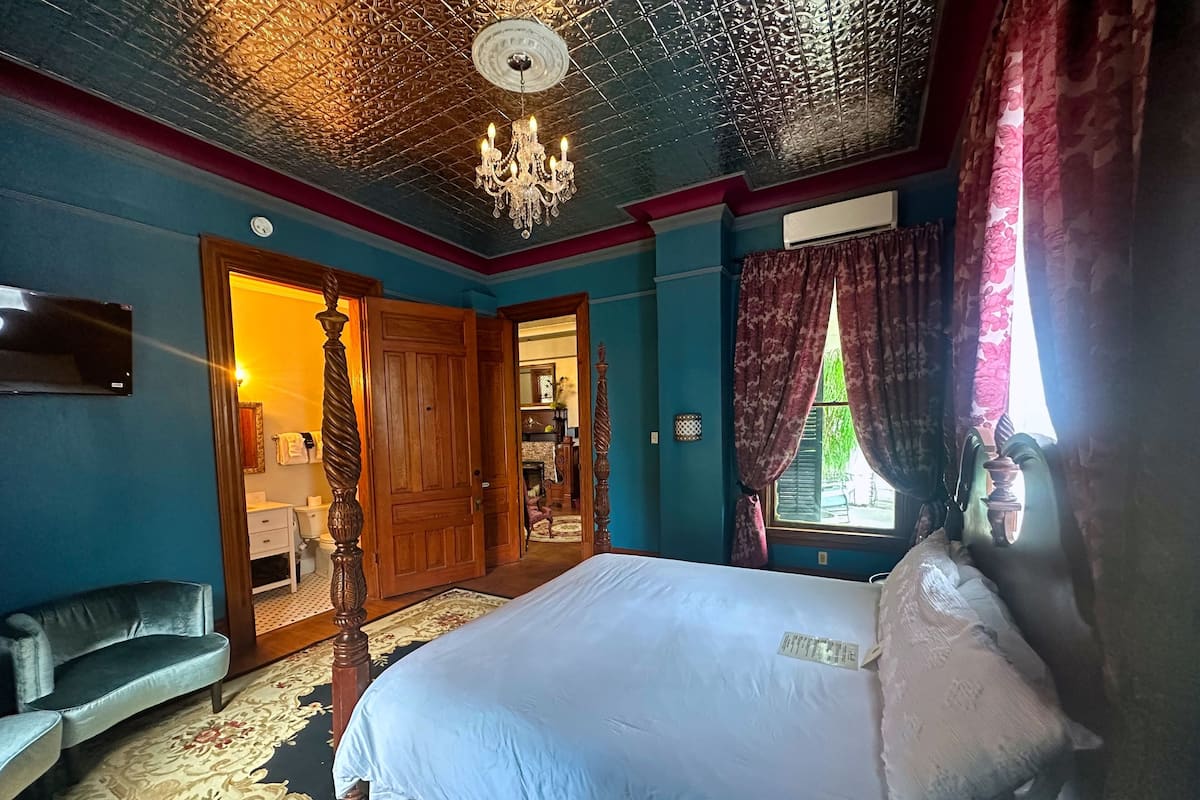 Une chambre à coucher avec un grand lit, un lustre, une armoire en bois et une vue sur une autre pièce par une porte ouverte.