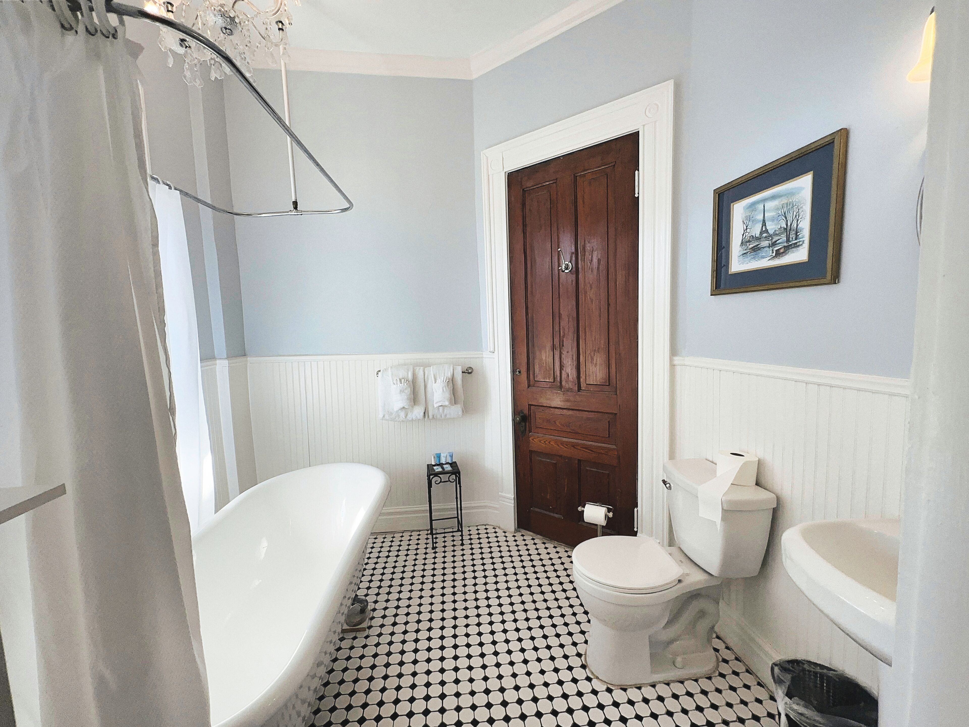 Une salle de bain avec une baignoire blanche, des toilettes et un lavabo. On y trouve une porte en bois et un tableau encadré accroché au mur.