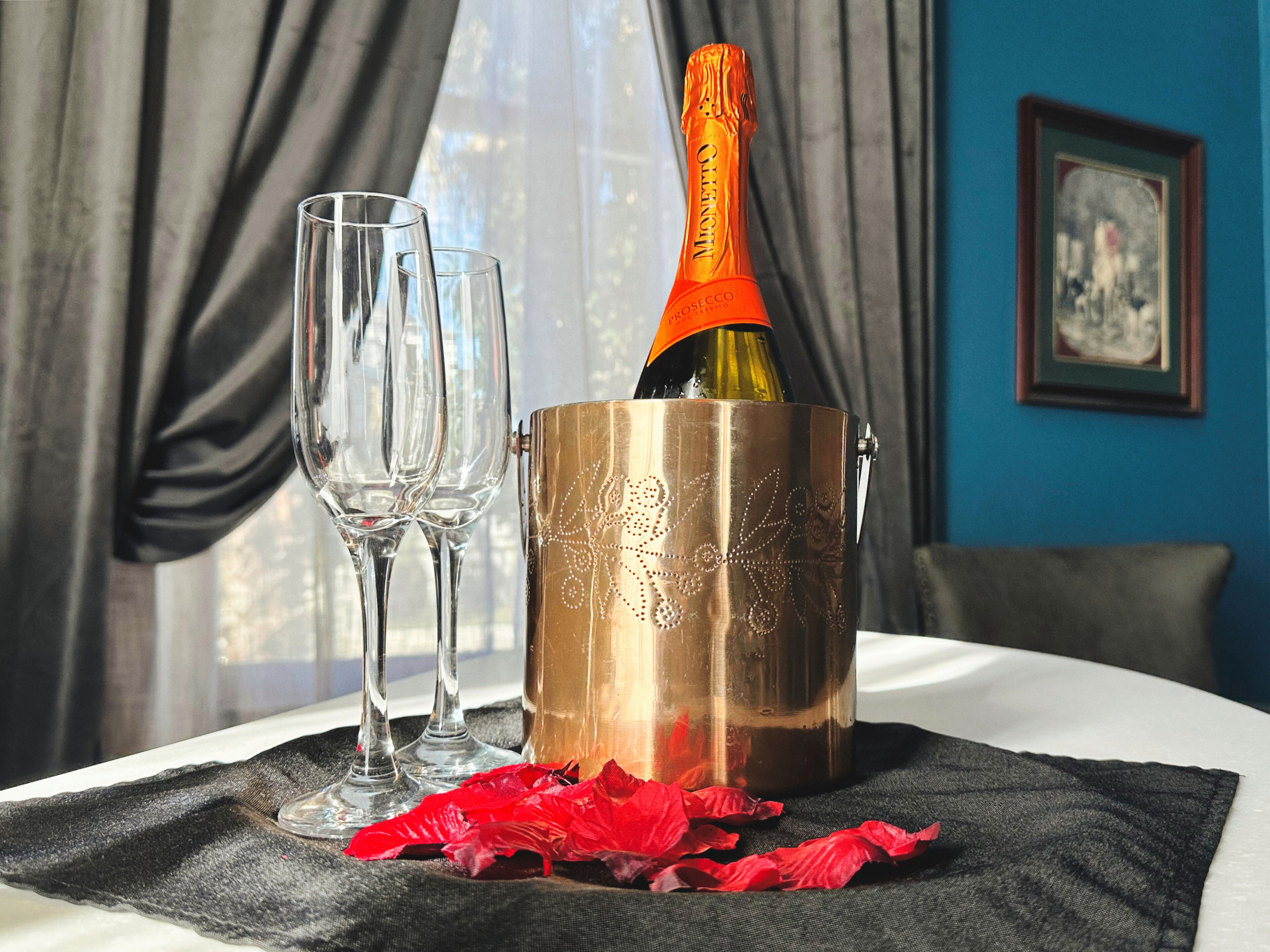 En champagneflaske i en metalspand med to glas ved siden af, placeret på et bord med et sort klæde og røde rosenblade.