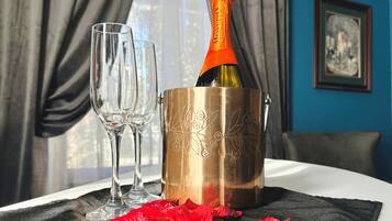 En champagneflaske i en metalspand med to glas ved siden af, placeret på et bord med et sort klæde og røde rosenblade.