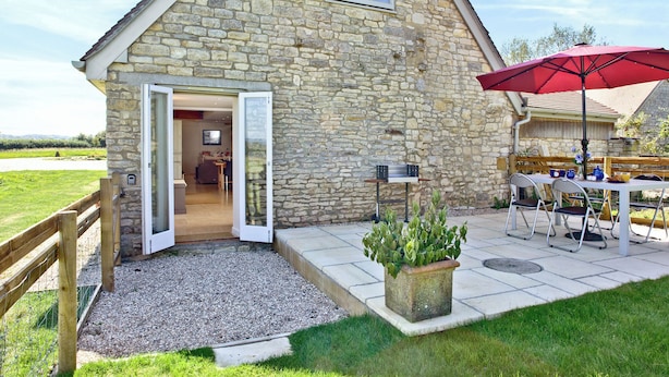 A stone building with a door open to a room, a table with chairs, a red umbrella, and a potted plant.