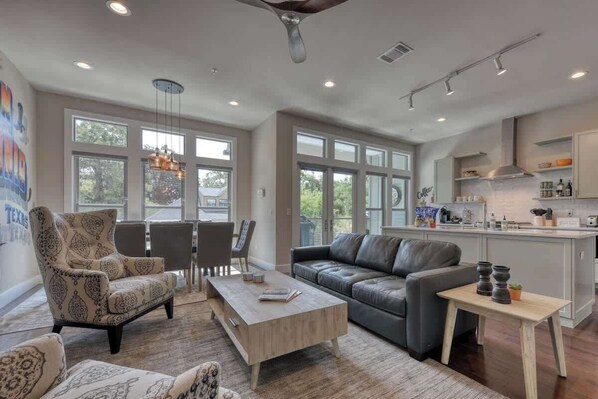 A spacious living room with a sofa, armchair, and coffee table, large windows, and a kitchen area with modern appliances.
