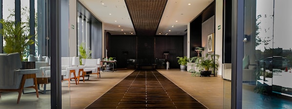 Un hall d’hôtel moderne avec un couloir central en bois foncé, des espaces de détente de part et d’autre, et des plantes en pot.