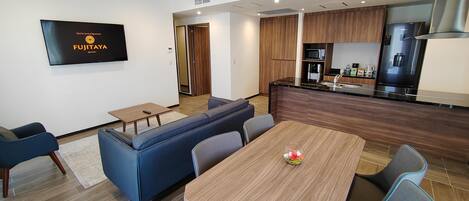 Una habitación de hotel moderna con zona de comedor, kitchenette y un televisor mostrando el nombre del hotel.