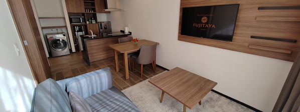 Ett kompakt hotellrum med ett litet pentry, en soffa, ett litet bord och en TV som är monterad på väggen.