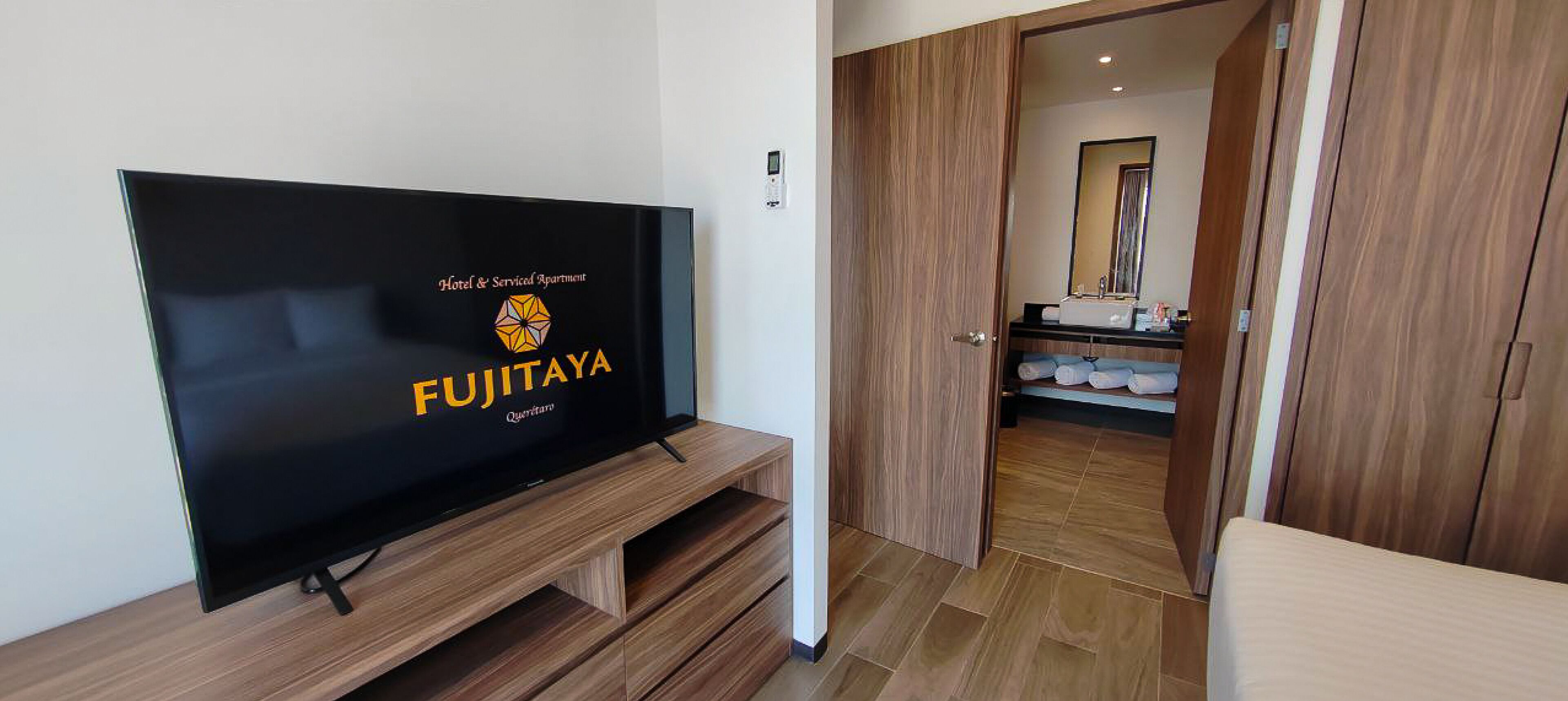 A hotel room with a flat-screen TV on a wooden stand, a wooden wardrobe, and a bathroom with a sink and mirror.
