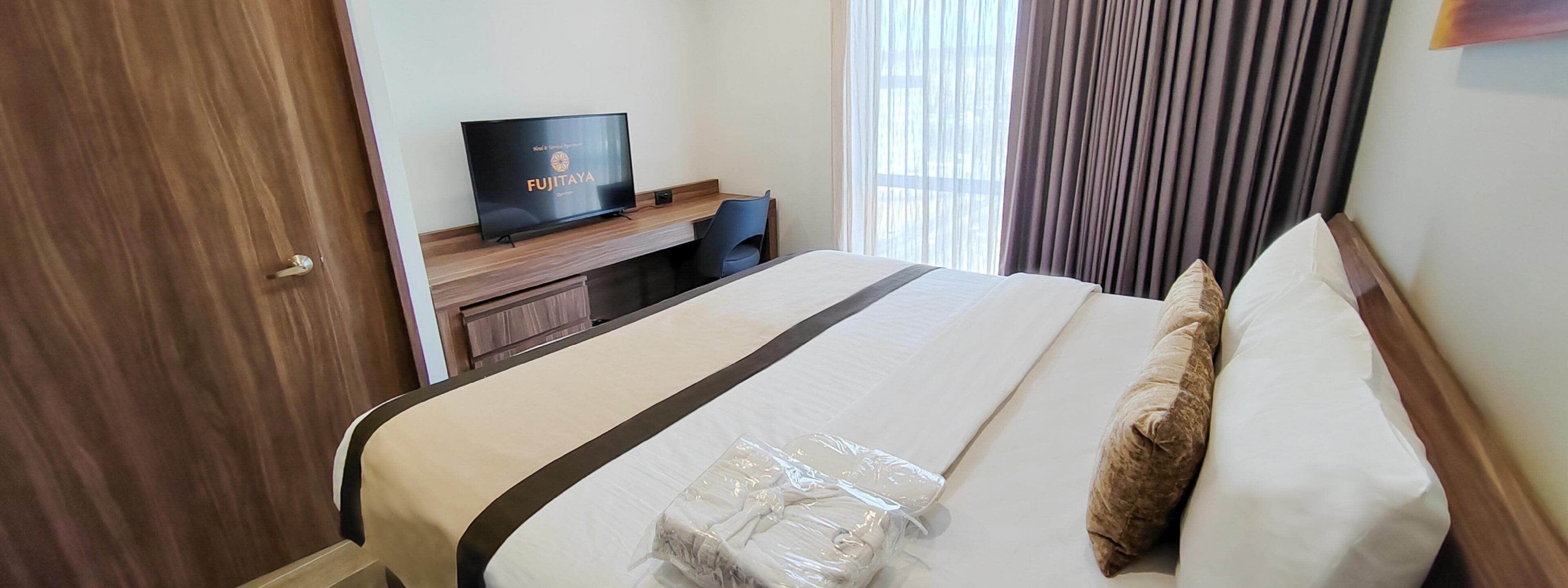 A hotel room with a bed, a TV on a wooden stand, a window with curtains, and a door.