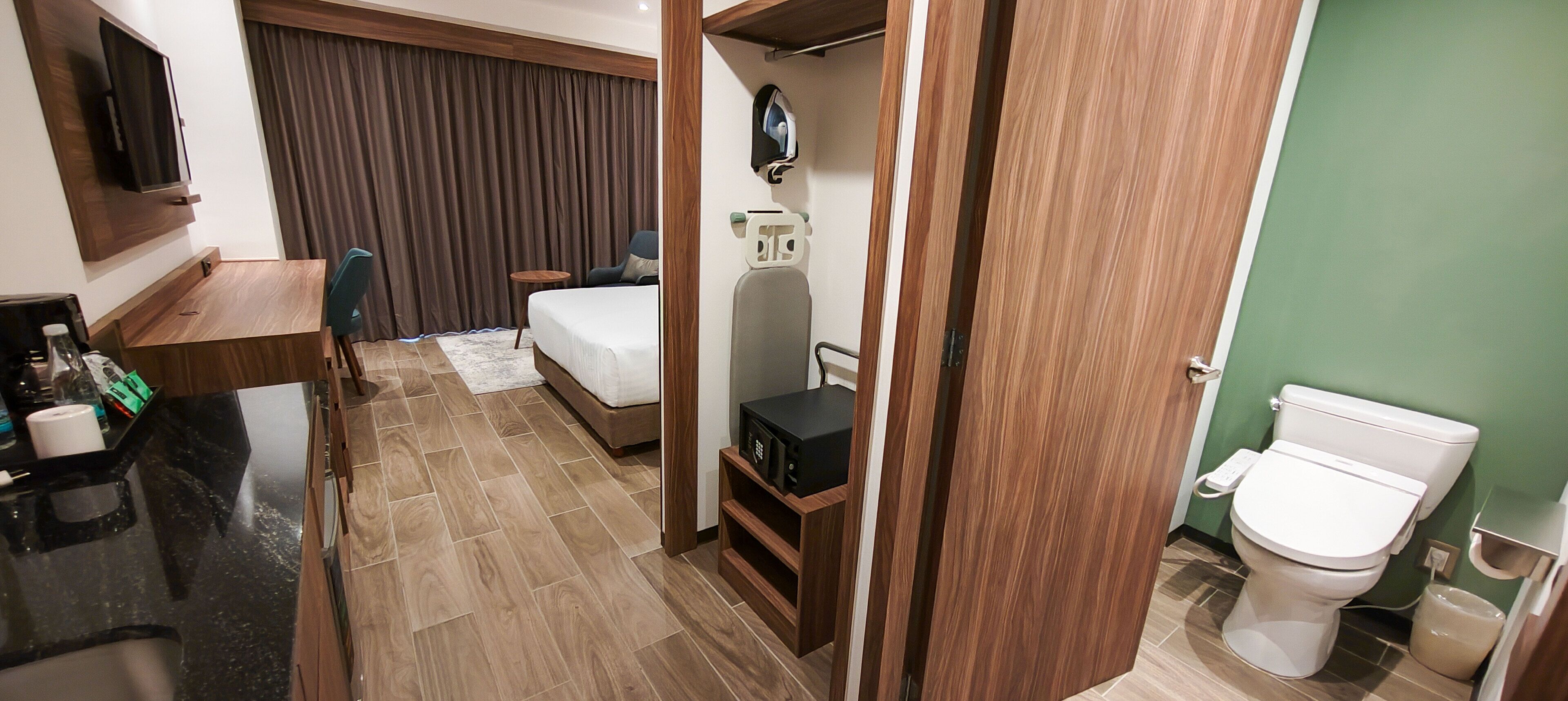 A hotel room with a bed, a desk, a TV, a small table, and a bathroom with a toilet and sink.