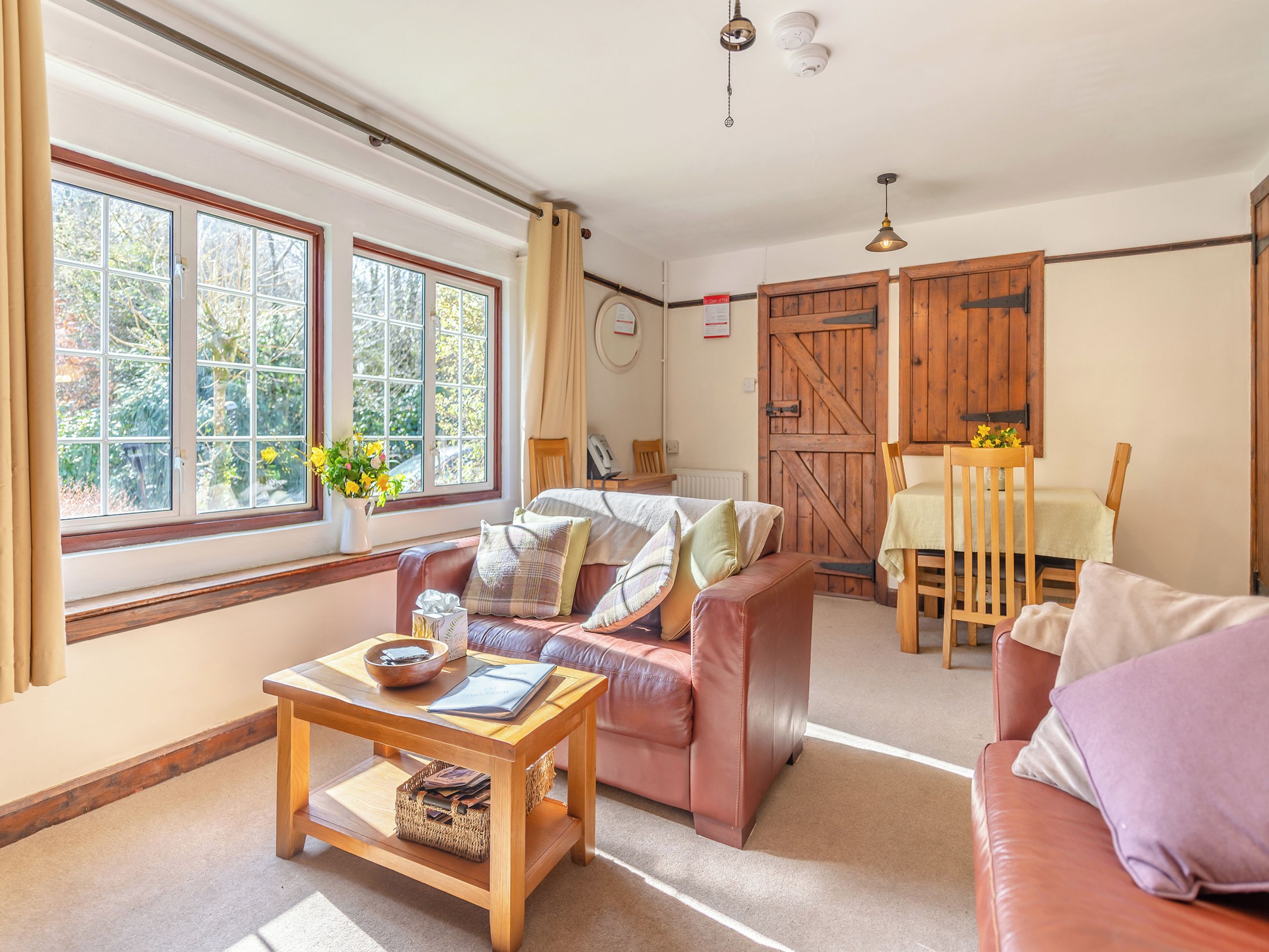 A cozy living room with a leather sofa, a wooden coffee table, and a dining table with chairs. Large windows allow natural light to fill the space.