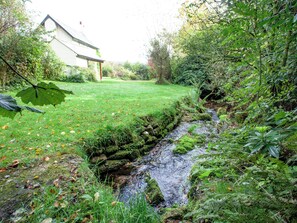 Ein kleiner Bach fließt durch eine Wiese, im Hintergrund sieht man ein Haus.