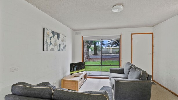 A modern living room with a grey sofa set, a wooden coffee table, and a flat-screen TV. A large window offers a view of a backyard with a lawn and trees. - Perfect for your beach holiday, you are only a stone's throw to the Port Fairy East Beach (Port Fairy)