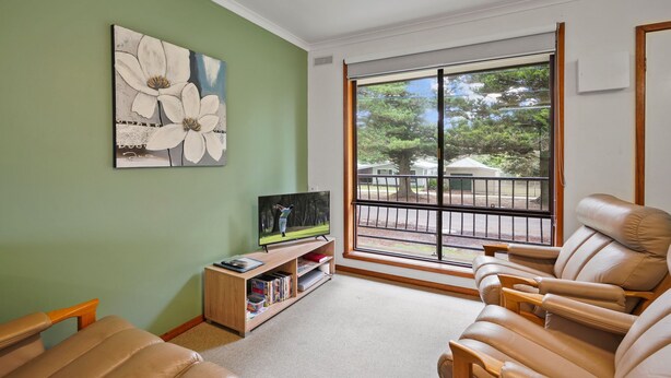 A living room with a television, a bookshelf, and a large window with a view of trees and a fence. - Perfect for your beach holiday, you are only a stone's throw to the Port Fairy East Beach (Port Fairy)