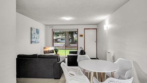 Une pièce de taille modeste, avec un canapé gris, une table blanche entourée de chaises, une grande fenêtre donnant sur une maison et des arbres, et une porte avec une plaque.
