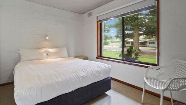 A bedroom with a bed, a nightstand, a window with a view of trees and a house, and a white chair. - Perfect for your beach holiday, you are only a stone's throw to the Port Fairy East Beach (Port Fairy)