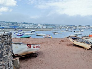 A coastal scene with boats moored on the sand and water, a stone wall, and a distant town.
