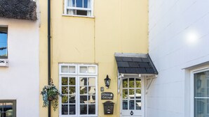 A yellow building with a white door, a window with a hanging flower basket, and a sign reading '13 Daisy Cottage.'