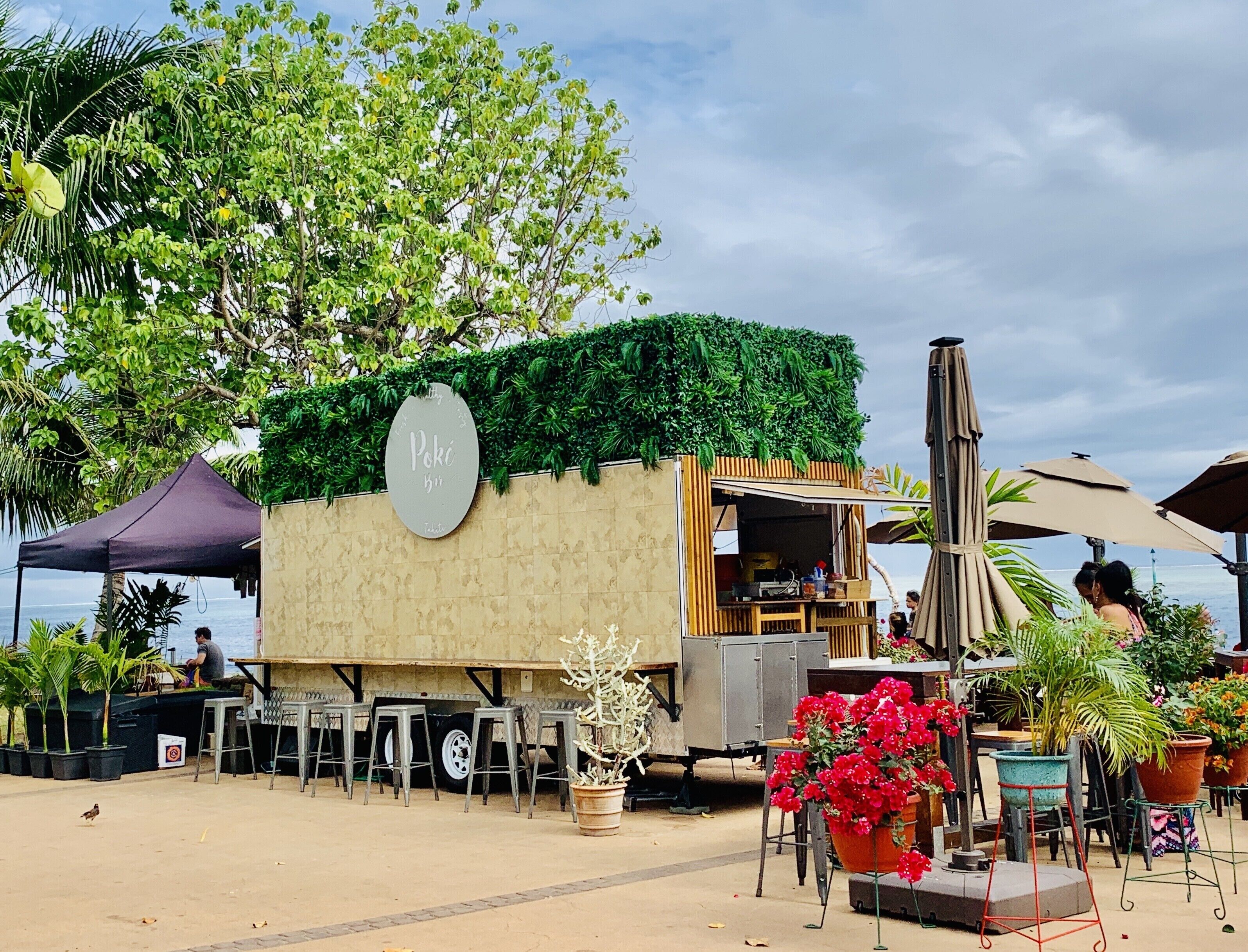 A food truck with a green roof and a sign that reads "Poke Bar". There are outdoor tables and chairs, and a few potted plants.