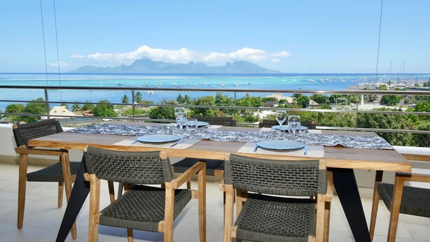 Un espace repas offrant une vue sur l’océan et les bateaux, avec une table en bois et des chaises en osier.
