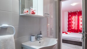 Een moderne badkamer met een witte wastafel, spiegel en handdoekhouder. Een slaapkamer met een bed en rode gordijnen is zichtbaar door een geopende deur.