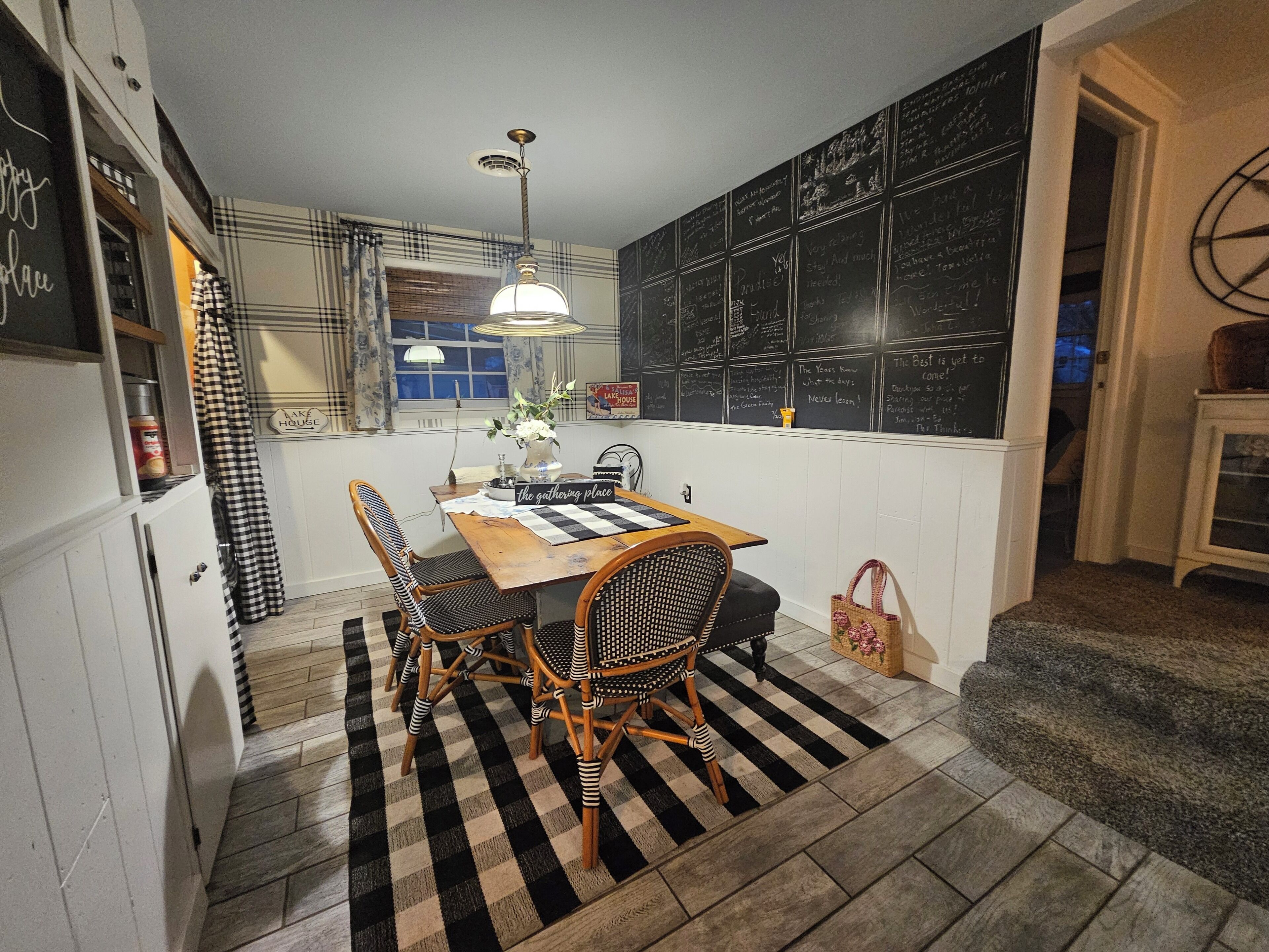 A dining area with a checkered rug, wooden table, wicker chairs, and a hanging light fixture.