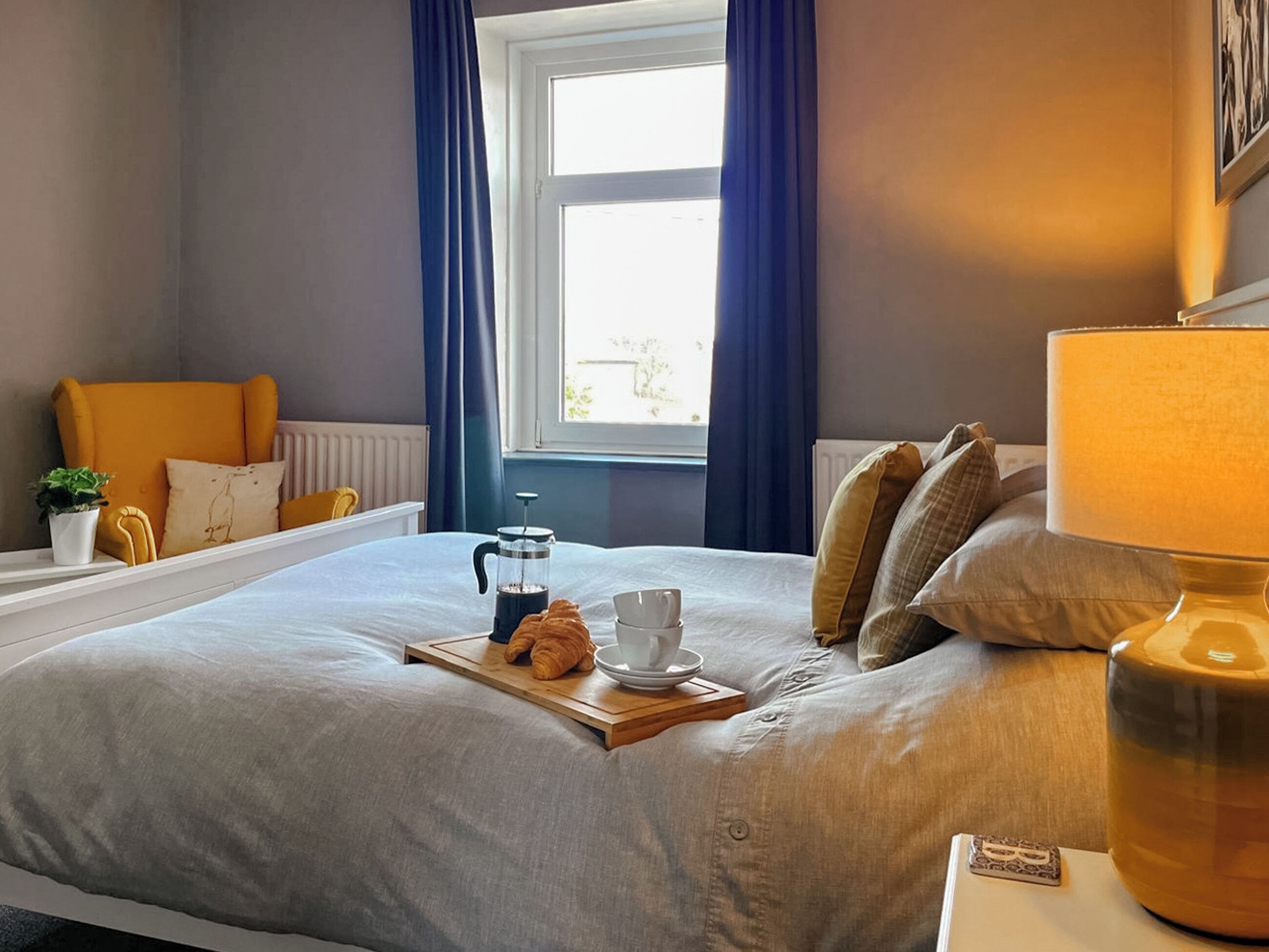 A neatly made bed with a tray of croissants and coffee, a bedside table with a lamp, and a window with blue curtains.