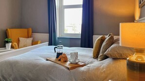 Un lit bien fait, avec un plateau contenant des croissants et du café, une table de chevet avec une lampe, et une fenêtre ornée de rideaux bleus.
