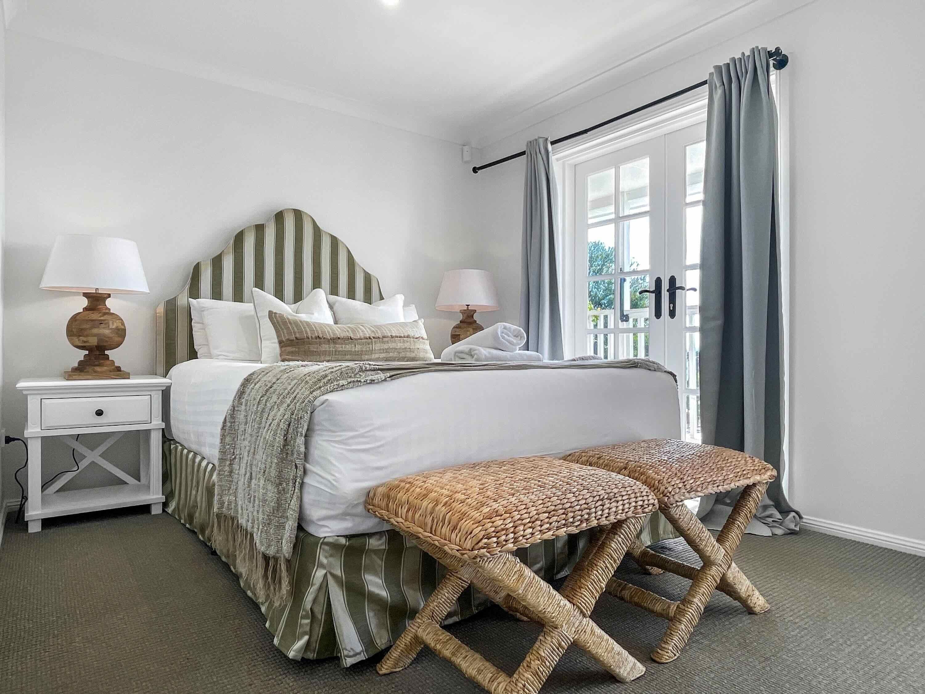 A bedroom with a large bed, two bedside tables with lamps, a window with curtains, and a woven bench.
