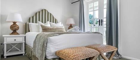 A bedroom with a large bed, two bedside tables with lamps, a window with curtains, and a woven bench.