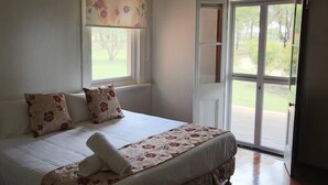 Une chambre à coucher avec un lit, des coussins à motifs floraux, une fenêtre donnant sur un paysage verdoyant et une porte-fenêtre coulissante.