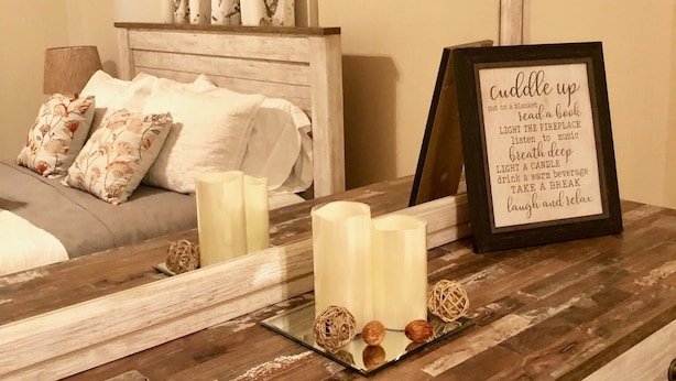 A cozy bedroom with a wooden headboard, a bedside table with candles, a framed sign, and a window with floral curtains.