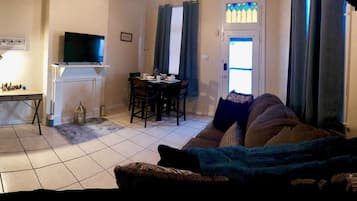 A living room with a TV on a stand, a dining table with chairs, and a sofa with a blanket.