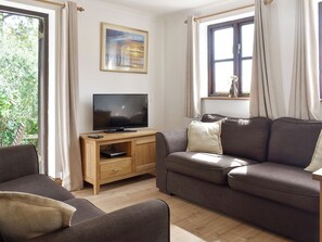 A living room with a flat-screen TV on a wooden stand, a brown sofa, and a window with curtains. - Prince Croft Cottage - UK12560 (Mount Hawke, near Redruth)