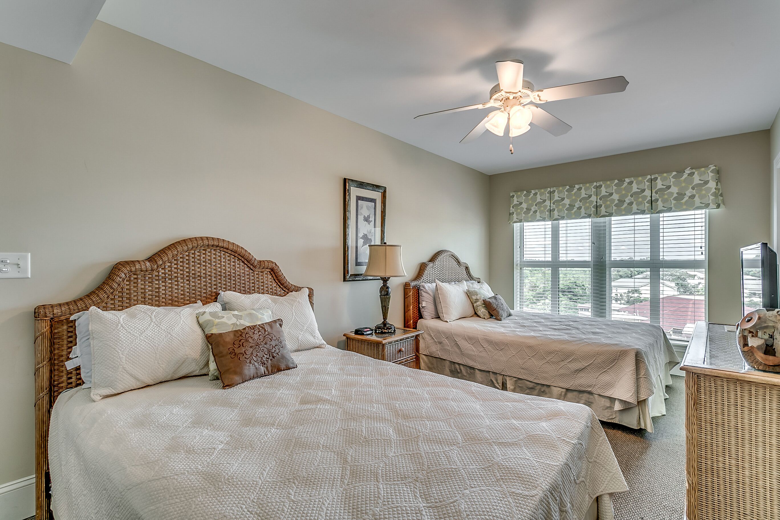 A bedroom with two beds, a ceiling fan, a lamp, and a large window with blinds.