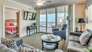 A spacious living area with a ceiling fan, a flat-screen TV, a balcony with a view, and a dining table with remote controls.