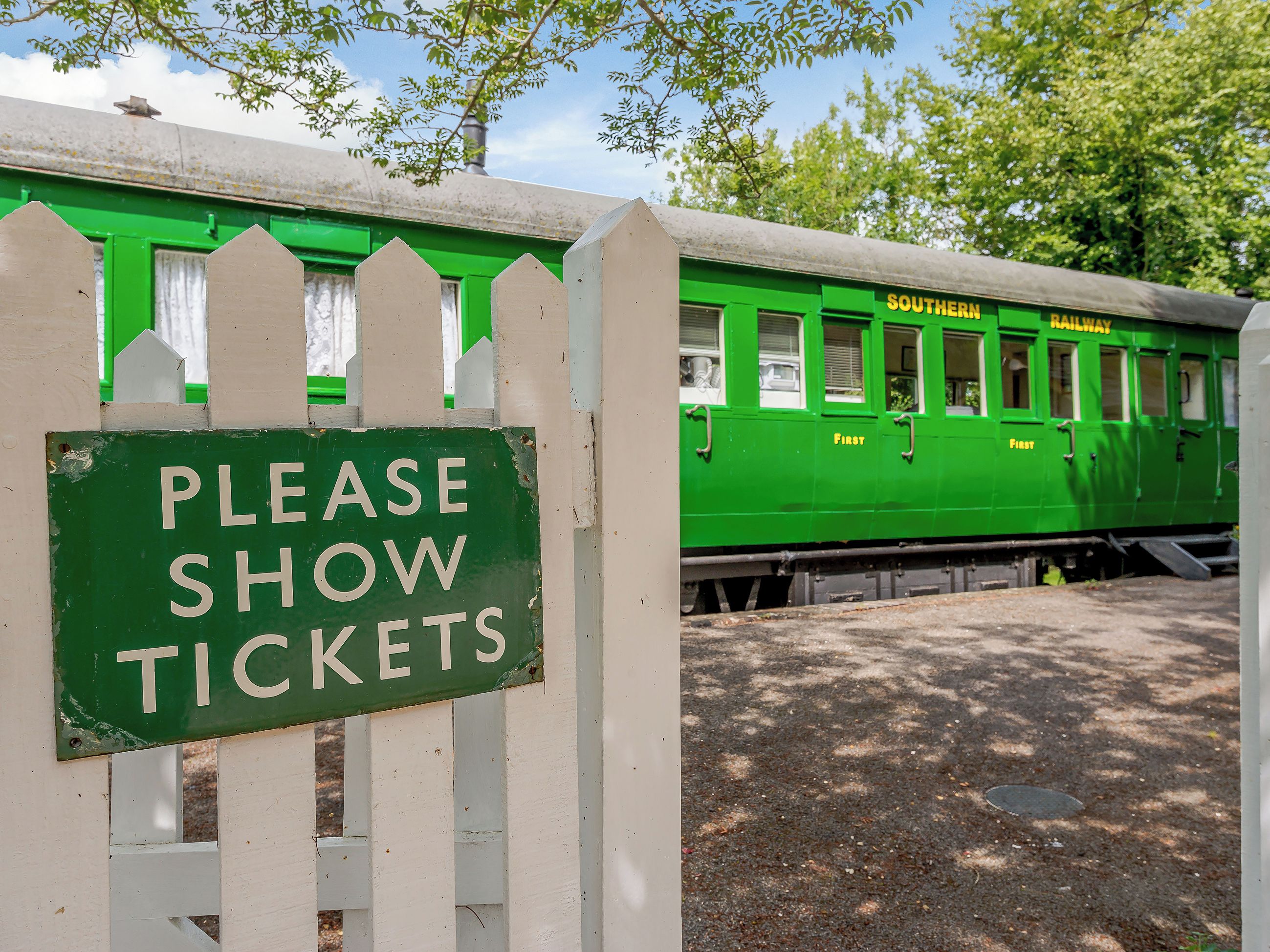 A green train with 'SOUTHERN RAILWAY' and 'FIRST' on it, passing by a white picket fence with a 'PLEASE SHOW TICKETS' sign.