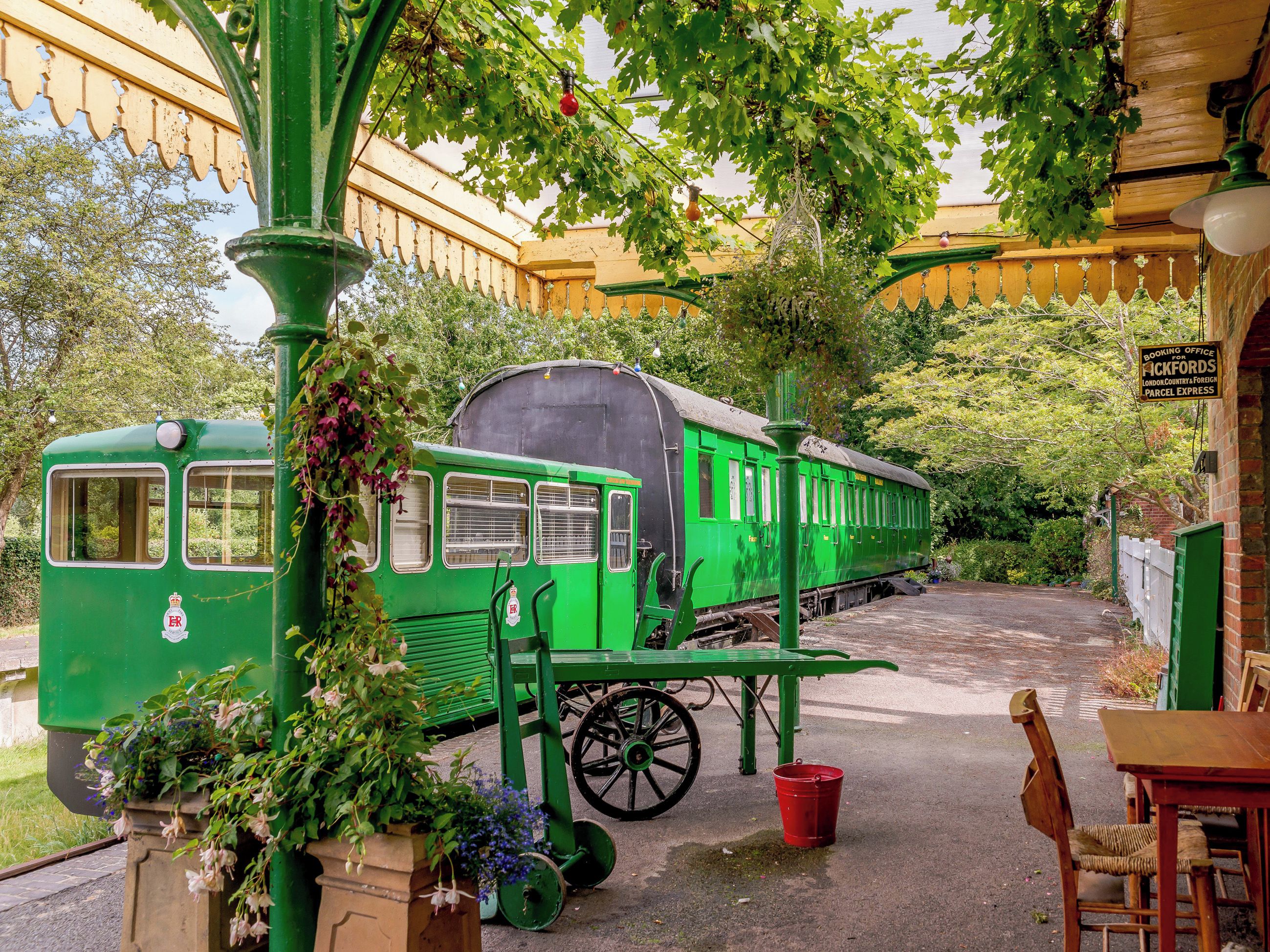 Un vagón de tren verde antiguo estacionado en un jardín, con un letrero de oficina de reservas, una mesa y sillas.