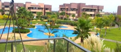 A balcony with a view of a swimming pool, palm trees, and a building complex.