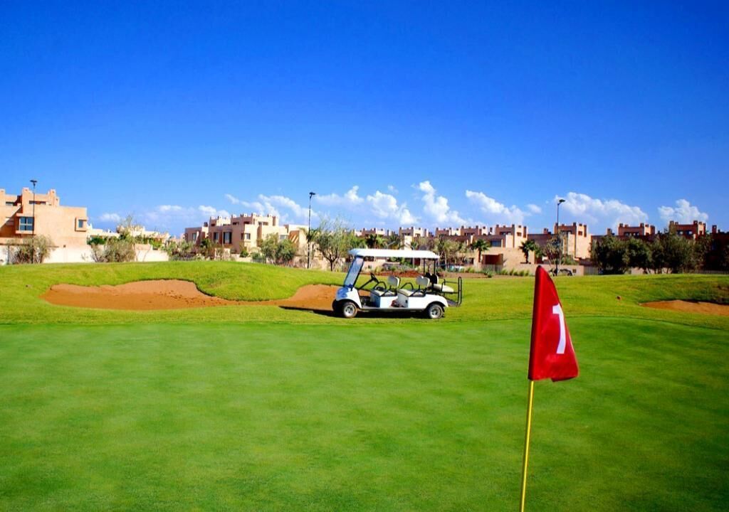 A golf course with a flag, a golf cart, and buildings in the background.