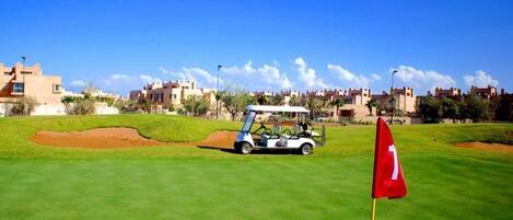 A golf course with a flag, a golf cart, and buildings in the background.