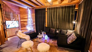 A cozy living room with a black sofa, white furry ottomans, a wooden TV stand, and a glass table with a candle. - Spacieux Chalet, proche des Télésièges,  Terrasse (Valloire)