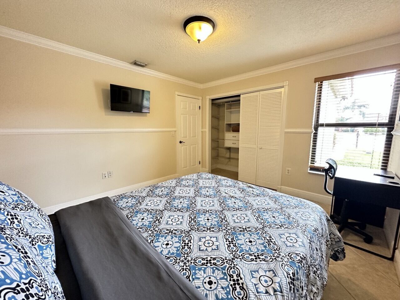 A bedroom with a bed, a desk, a chair, a TV, a closet, and a window.