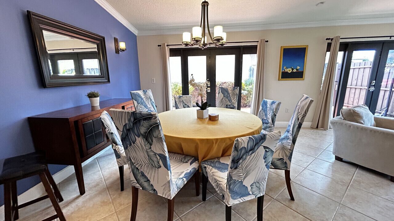 A dining area with a round table, chairs with a tropical leaf pattern, a chandelier, a wooden sideboard, and a sofa.