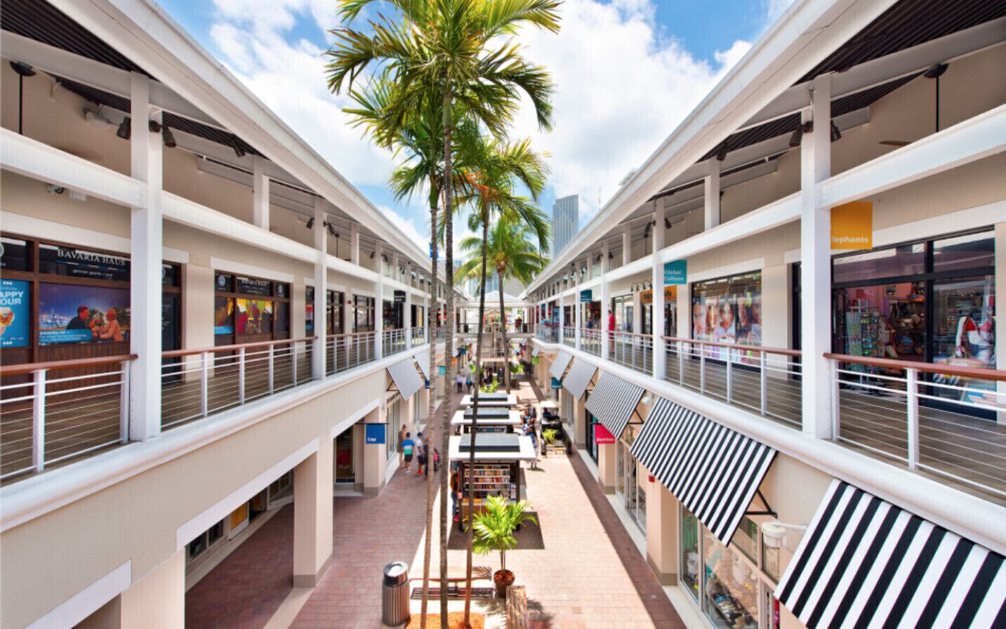 A multi-level shopping mall with a central walkway, palm trees, and various retail stores.