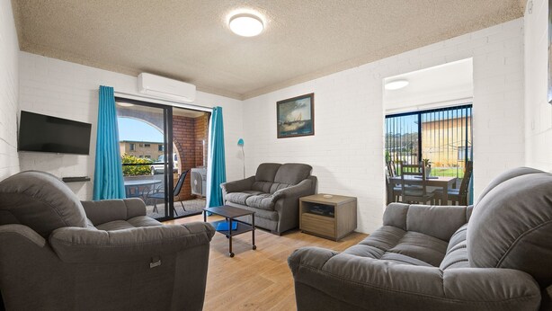 A modern living room with grey sofas, a flat-screen TV, and a dining area with a table and chairs. - Apollo Unit 17 - Ground Floor (Narooma)