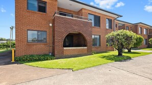 A modern brick building with a central arched window, a small garden, and a paved driveway. - Apollo Unit 17 - Ground Floor (Narooma)